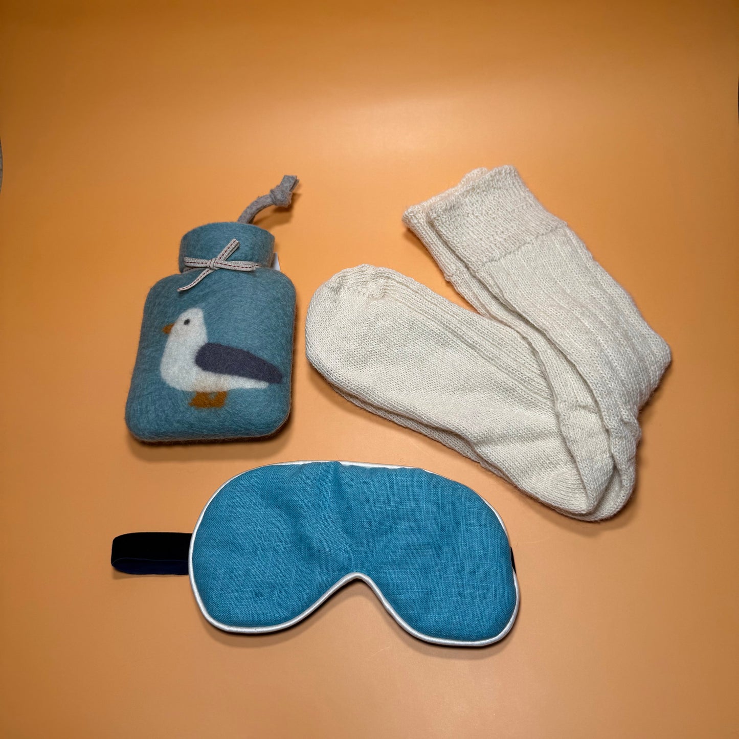 Teal seagull motif mini hotwater bottle, ivory Angora bed socks and blue linen eye mask. All on a soft orange background.