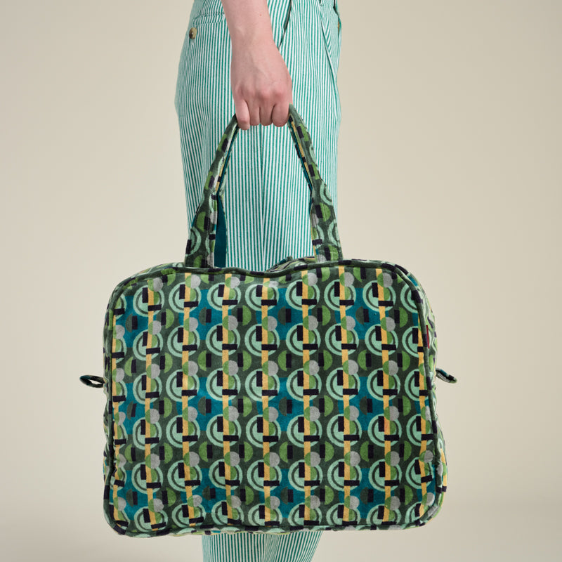 A person wearing mint green striped trousers and black slippers holds a large zippered travel bag with an allover geometric chain-link pattern in green, teal, yellow, and black against a plain beige studio background.