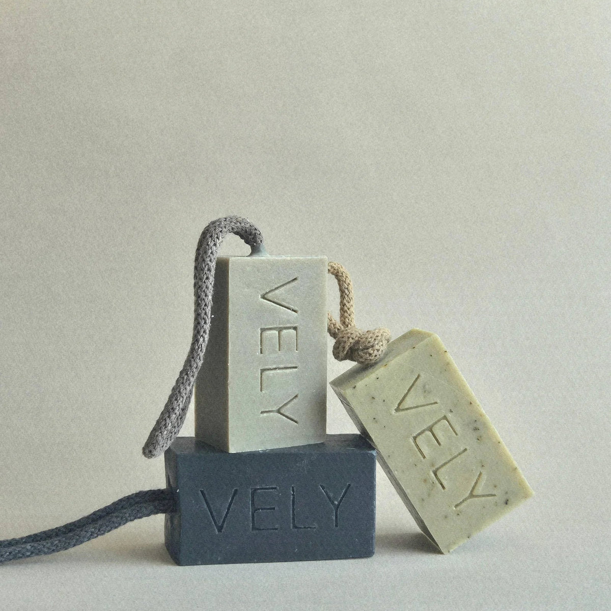 Three rectangular soap bars labeled VELY, each with a cord loop, arranged on a plain light background. One soap is dark charcoal gray, and two are pale warm gray-beige.