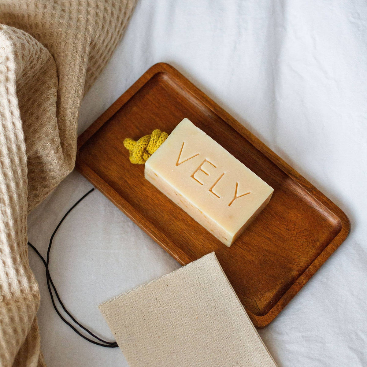 Bar of VELY soap on a wooden tray with a matching soap bag on a white background