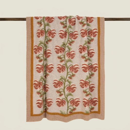 A woven textile panel, a wool trhow, featuring a vertical floral pattern with terracotta flowers, green stems, and a mustard-gold border on a light beige background, hanging from a dark wooden rod.