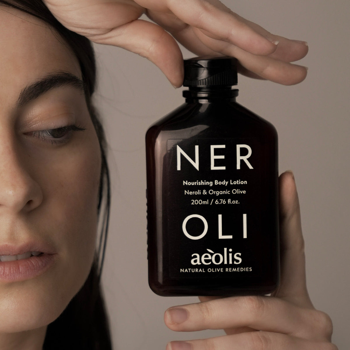 A close-up lifestyle shot of a woman holding a dark amber bottle of aloe vera body lotion. The bottle features white text reading 'NER OLI aeolis' and is held against a neutral, warm beige background.