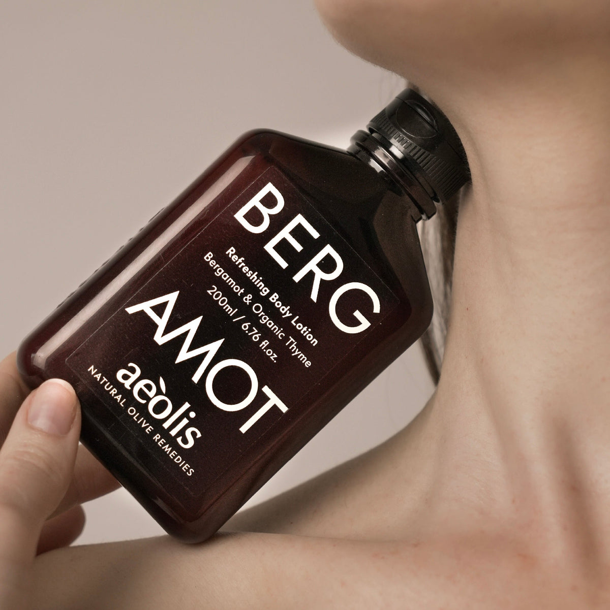 A hand holds a dark amber bottle of Berg Amot aeolis Natural Olive Beanchies Refreshing Body Lotion against a model's neck and shoulder, with text overlay at the bottom.