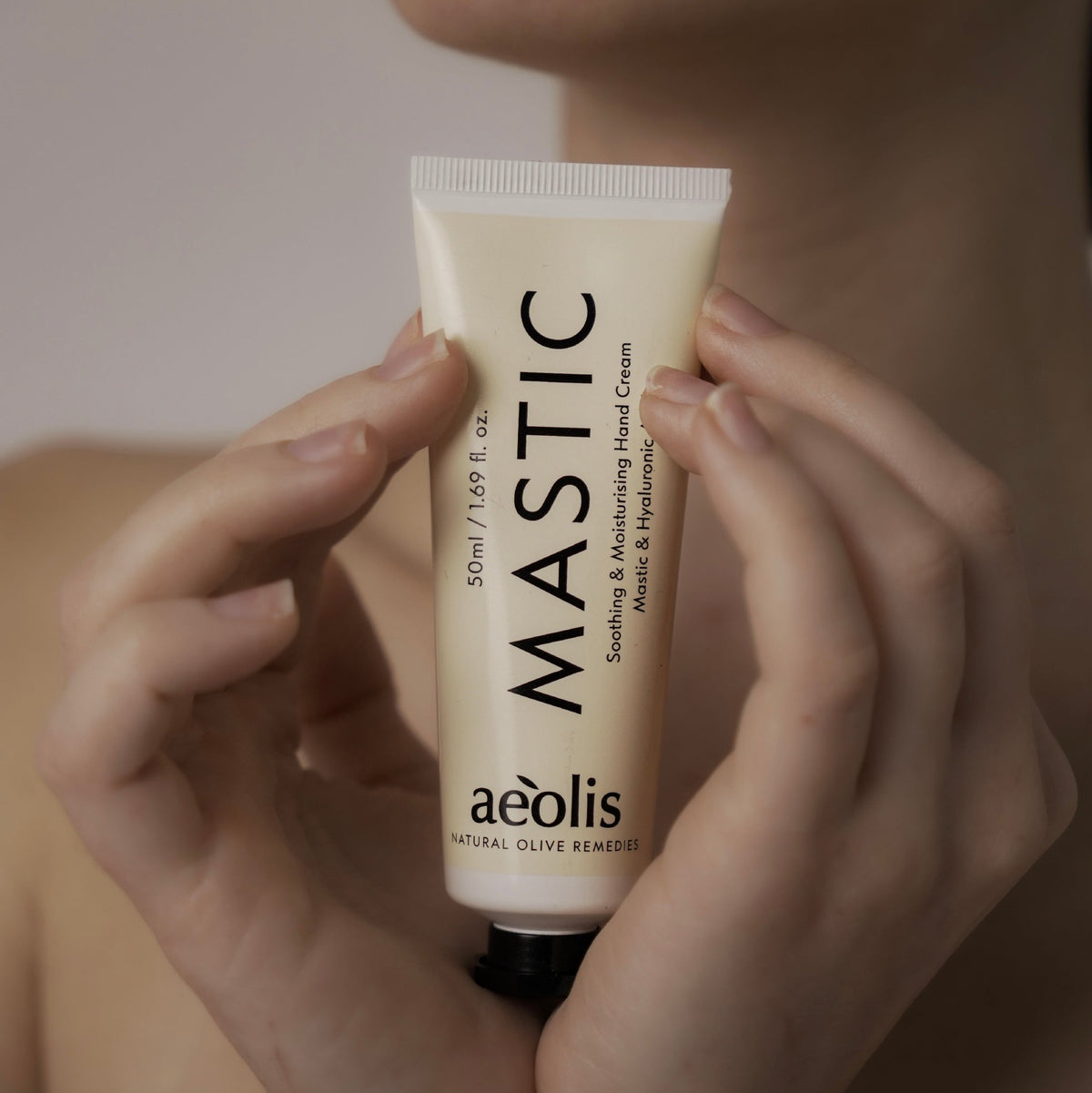 A person holds a squeeze tube of aeolis MASTIC Soothing & Moisturizing Hand Cream against their chest. The tube is a warm cream color with black text, and the background is a soft, warm beige.