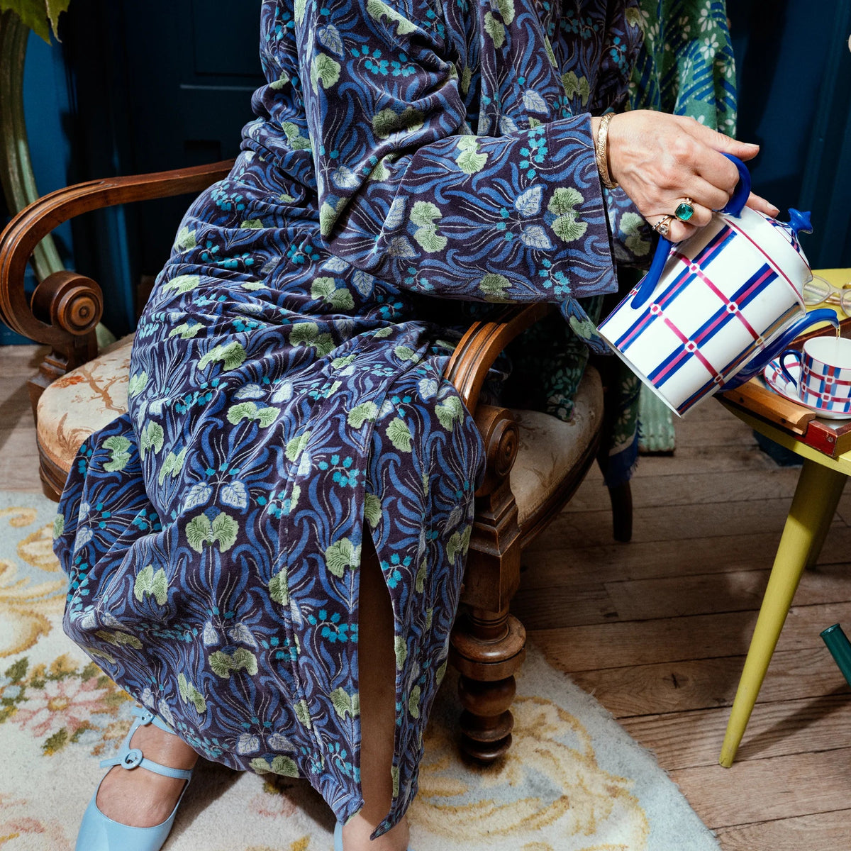 A person seated in a wooden armchair pours tea from a blue-and-white checkered teapot into a small cup on a yellow side table. The person is wearing a long blue floral robe, and the scene includes a vase of dried flowers, a green watering can, and a colorful fabric accessory on the table.