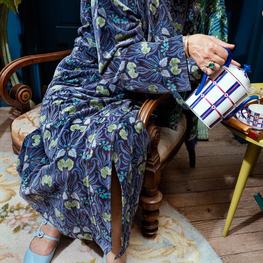 A person seated in a wooden armchair pours tea from a blue-and-white checkered teapot into a small cup on a yellow side table. The person is wearing a long blue floral robe, and the scene includes a vase of dried flowers, a green watering can, and a colorful fabric accessory on the table.