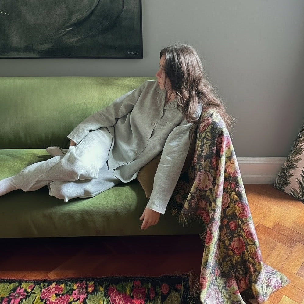 Woman sitting on a green couch with a floral blanket, surrounded by plants and a painting.