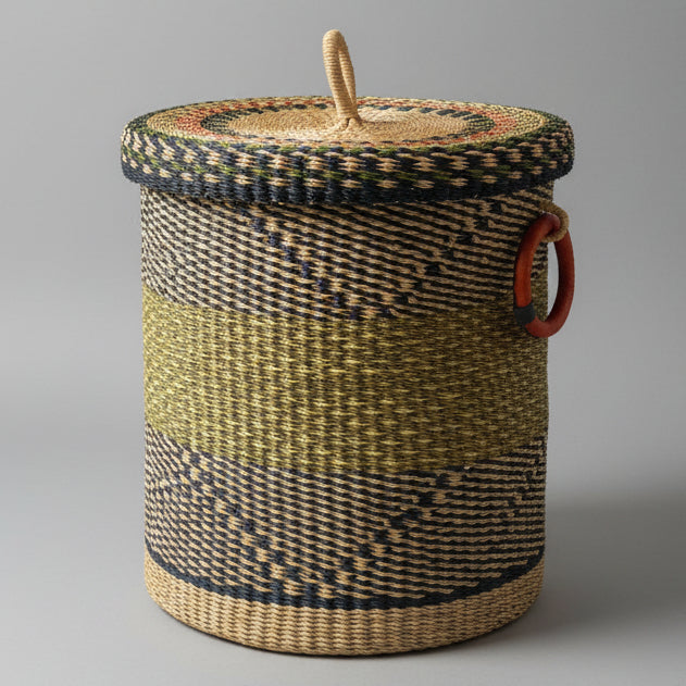 A large woven basket with a lid, featuring a natural tan body with dark brown and black patterned bands, a wooden handle on top, and a red hanging loop on the side, set on a wooden floor.