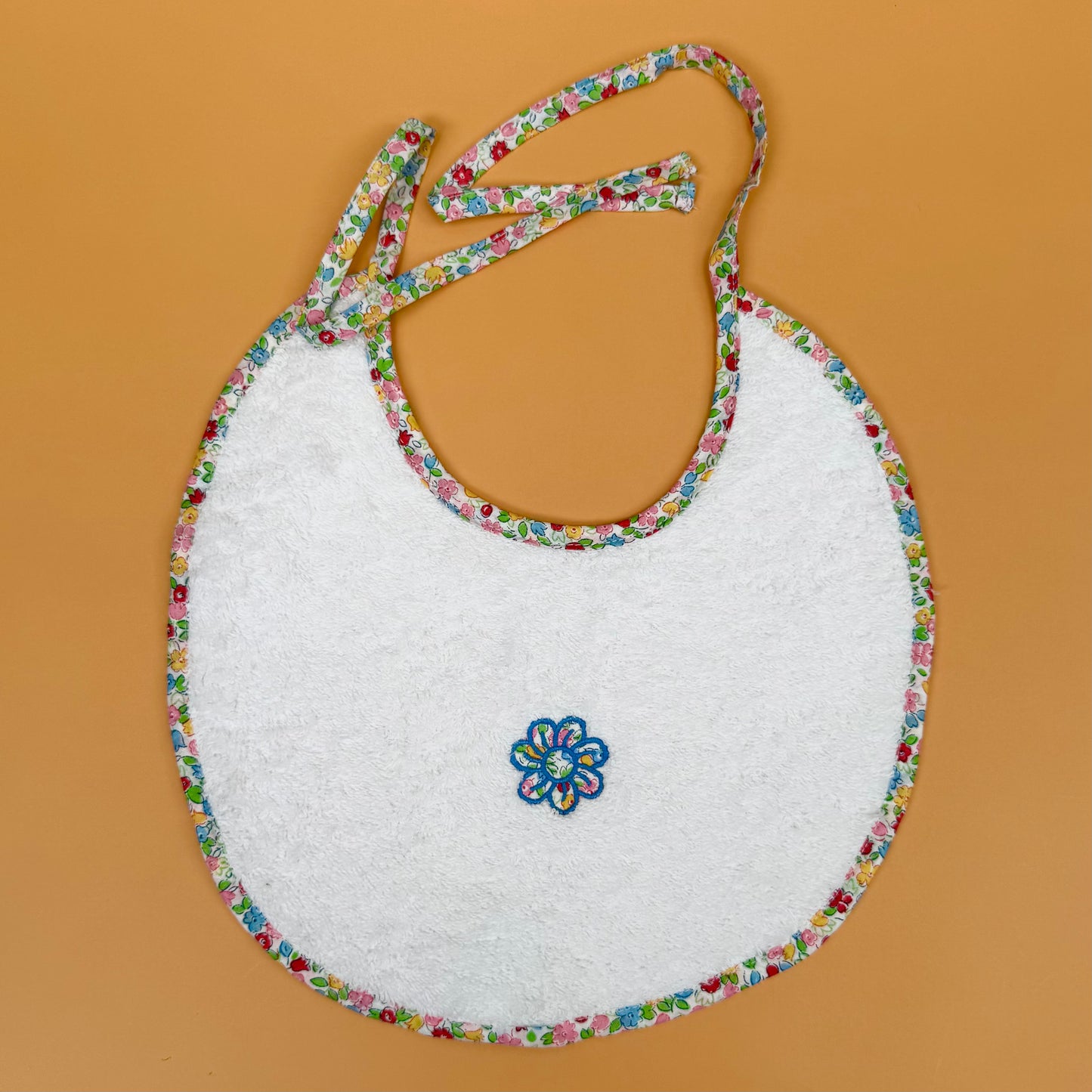 White bib with colorful floral trim and embroidered flower on an orange background