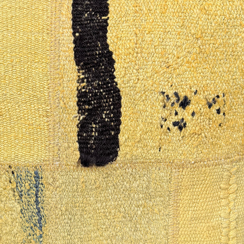 A square yellow woven throw pillow with a vertical black stripe and distressed details, resting on a weathered wooden bench against a dark green wall.