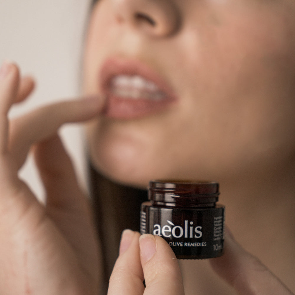 A close-up lifestyle shot of a person holding a small amber glass jar of aeolis Olive Remedies while applying a cream to their lips.