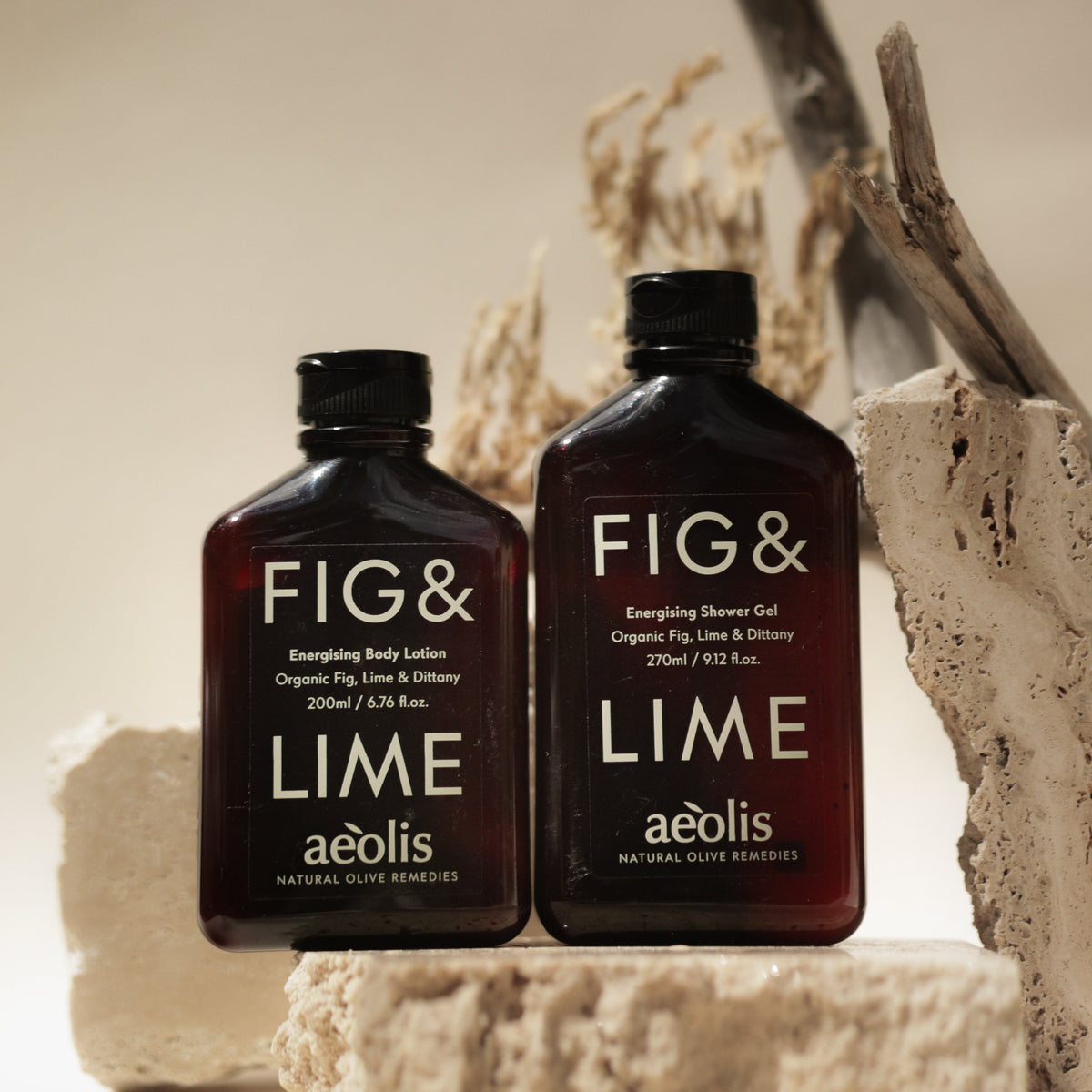 Two dark amber bottles of aólis Fig & Lime body and skin care products, shown on a beige stone prop with dried branches in the background.
