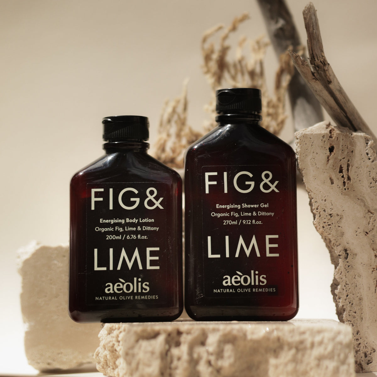 Two dark amber bottles of aólis Fig & Lime body and skin care products, shown on a beige stone prop with dried branches in the background.