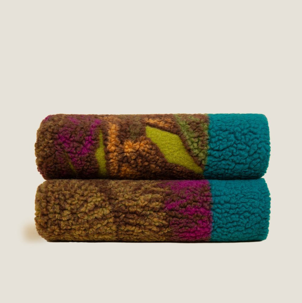 Two folded textured sherpa blankets stacked vertically, featuring a multicolor abstract pattern with brown, olive green, magenta, and teal sections, set against a solid light beige background.
