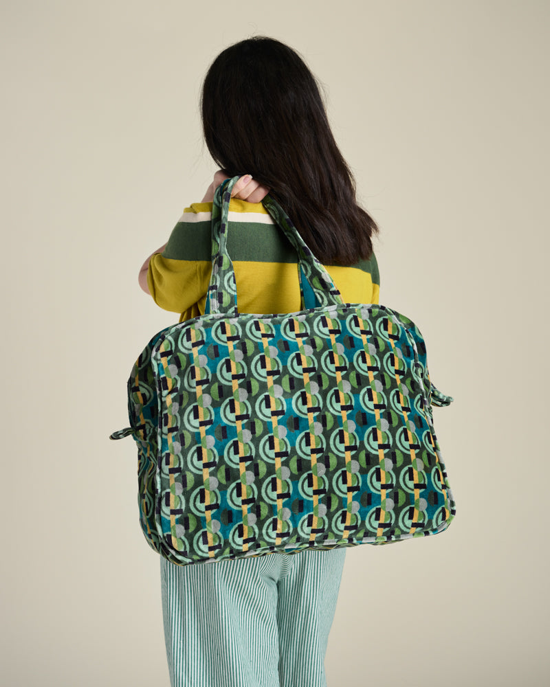 A woman viewed from behind carries a large patterned duffel bag over her shoulder. The bag has an allover geometric circular print in teal, green, yellow, and white.
