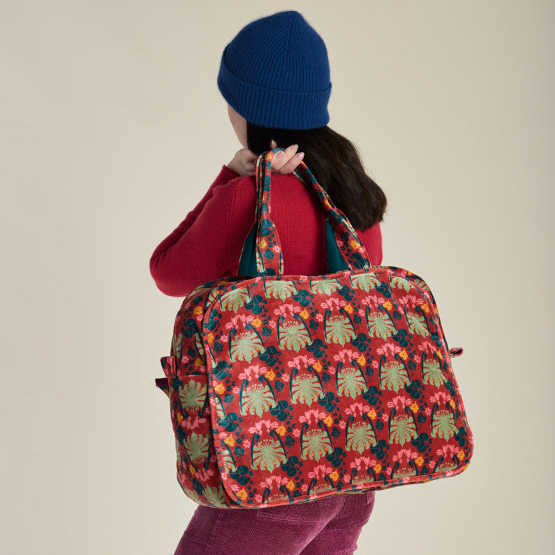 A person wearing a blue beanie, red top, and magenta pants carries a large duffel bag with a colorful tropical floral and palm leaf pattern, photographed against a plain beige studio background.
