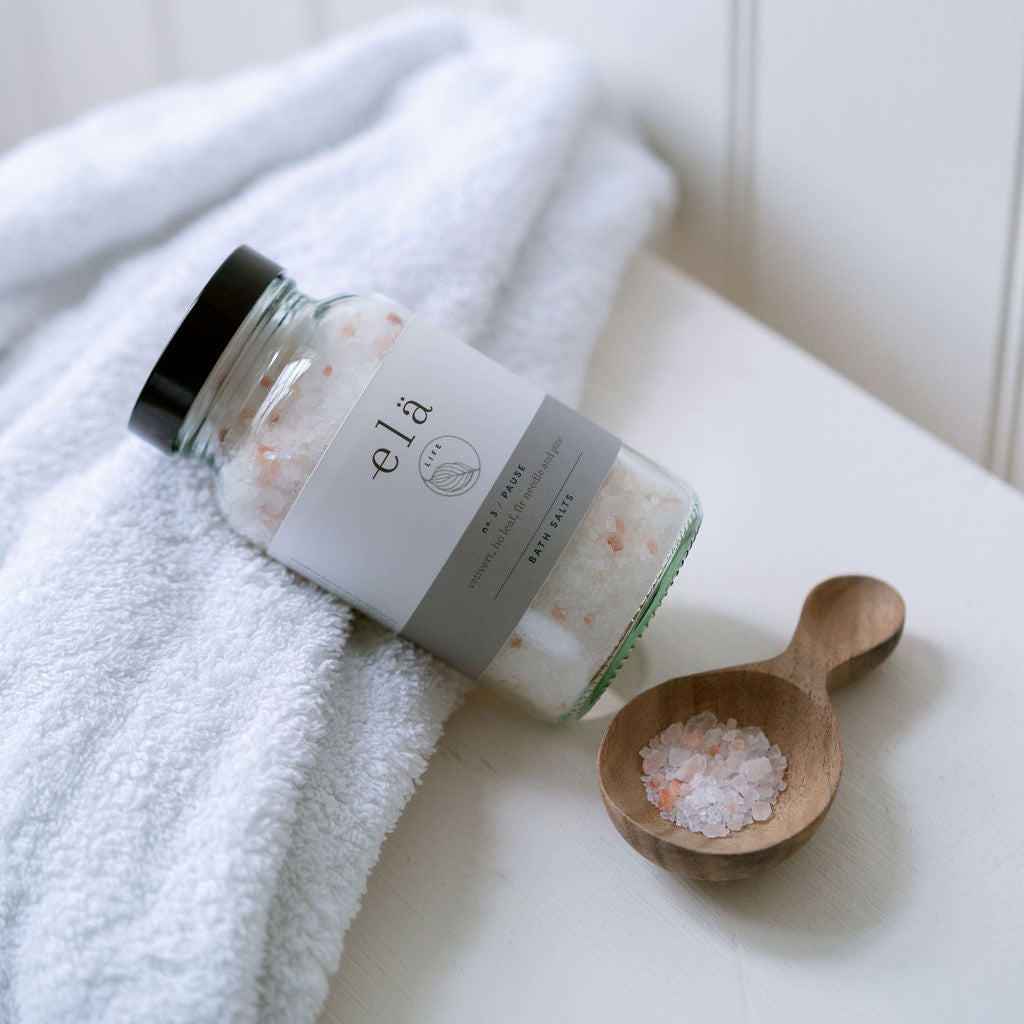 Bottle of elá bath salts with a wooden scoop on a white surface.