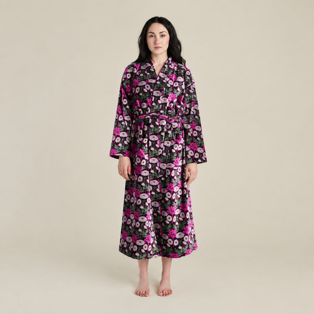A woman modeling a long-sleeved floral robe with a matching waist tie, featuring a black background with pink, purple, and white flowers, against a solid light beige studio backdrop.