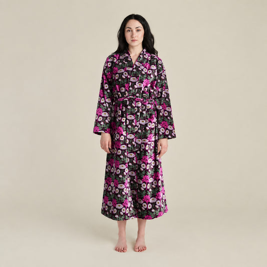 A woman modeling a long-sleeved floral robe with a matching waist tie, featuring a black background with pink, purple, and white flowers, against a solid light beige studio backdrop.