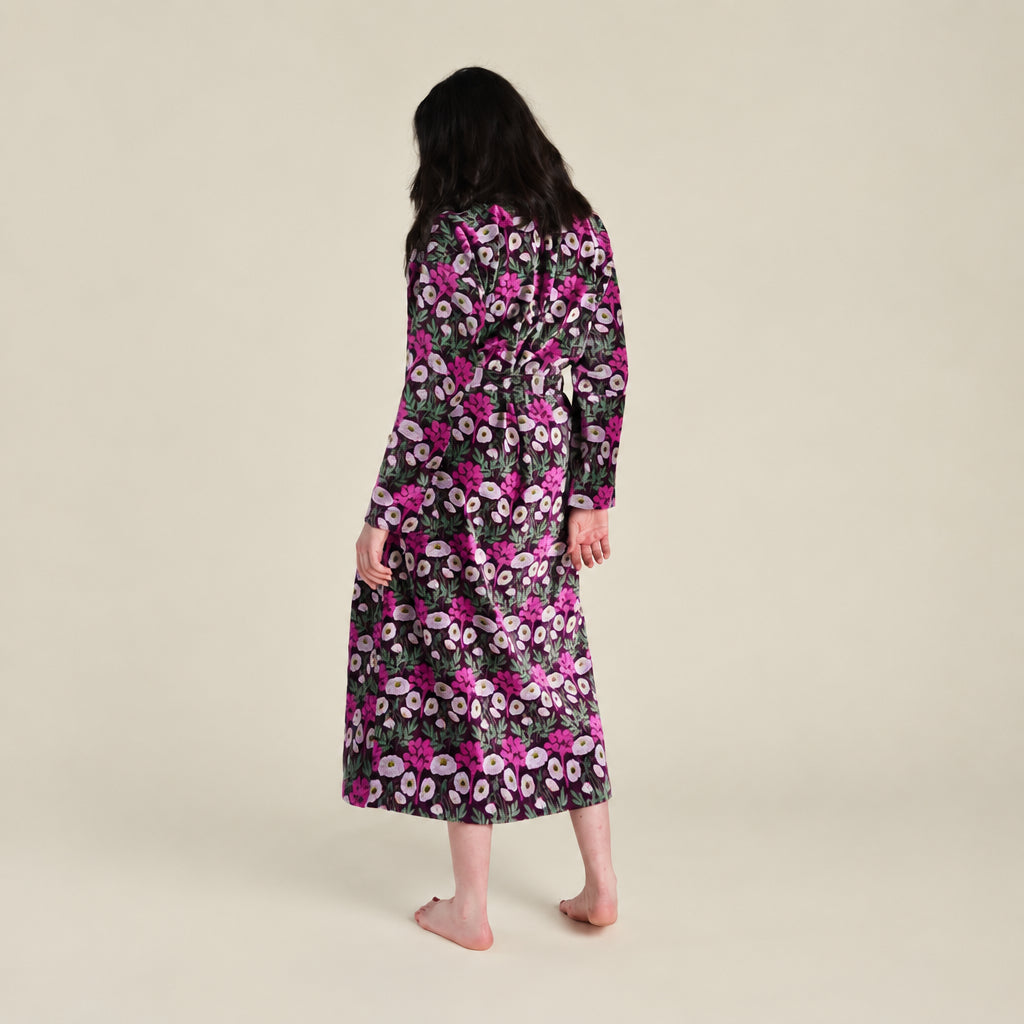 A woman stands barefoot against a plain beige studio background, photographed from the back while wearing a long-sleeve maxi robe with a black base and a dense pink, purple, and green floral pattern.