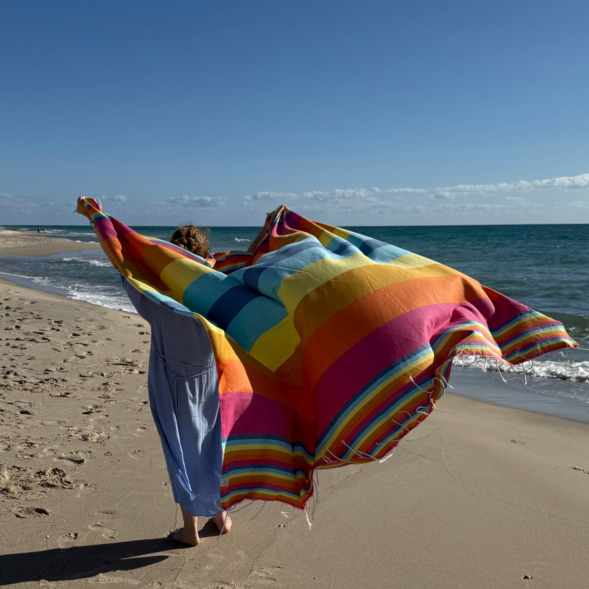 Rainbow beach towel being held up on a beach, blowing in the wind.