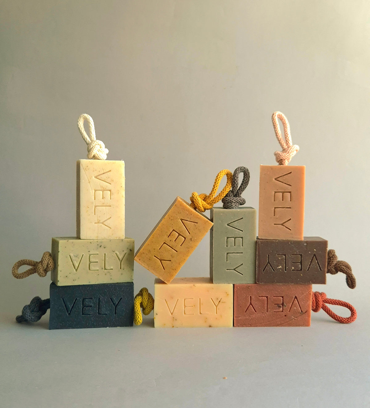 Six rectangular soap bars in muted earthy tones, each embossed with the brand name 'VELY' and tied with a small rope loop, arranged in small stacks on a neutral studio background.