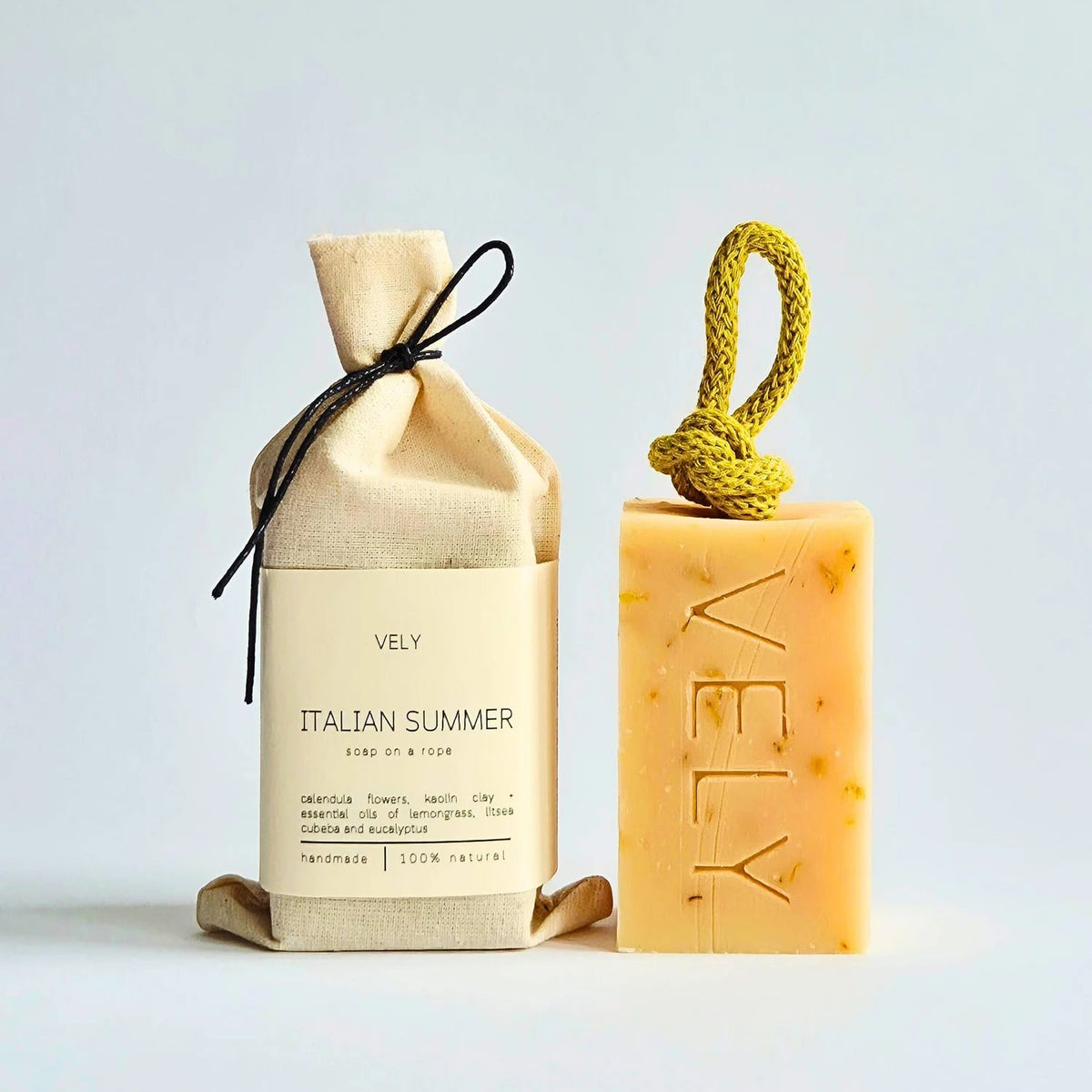 A handmade bar of soap and its fabric pouch packaging on a light gray background. The soap is a warm yellow-orange with visible botanical inclusions, featuring a yellow cord loop and the brand name 'VELY' embossed on the front.